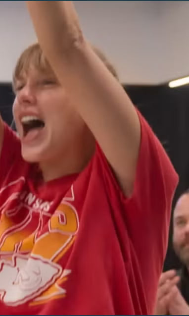 Taylor Swift in a JUNKFOOD red shirt with a graphic design, arms raised in an indoor setting