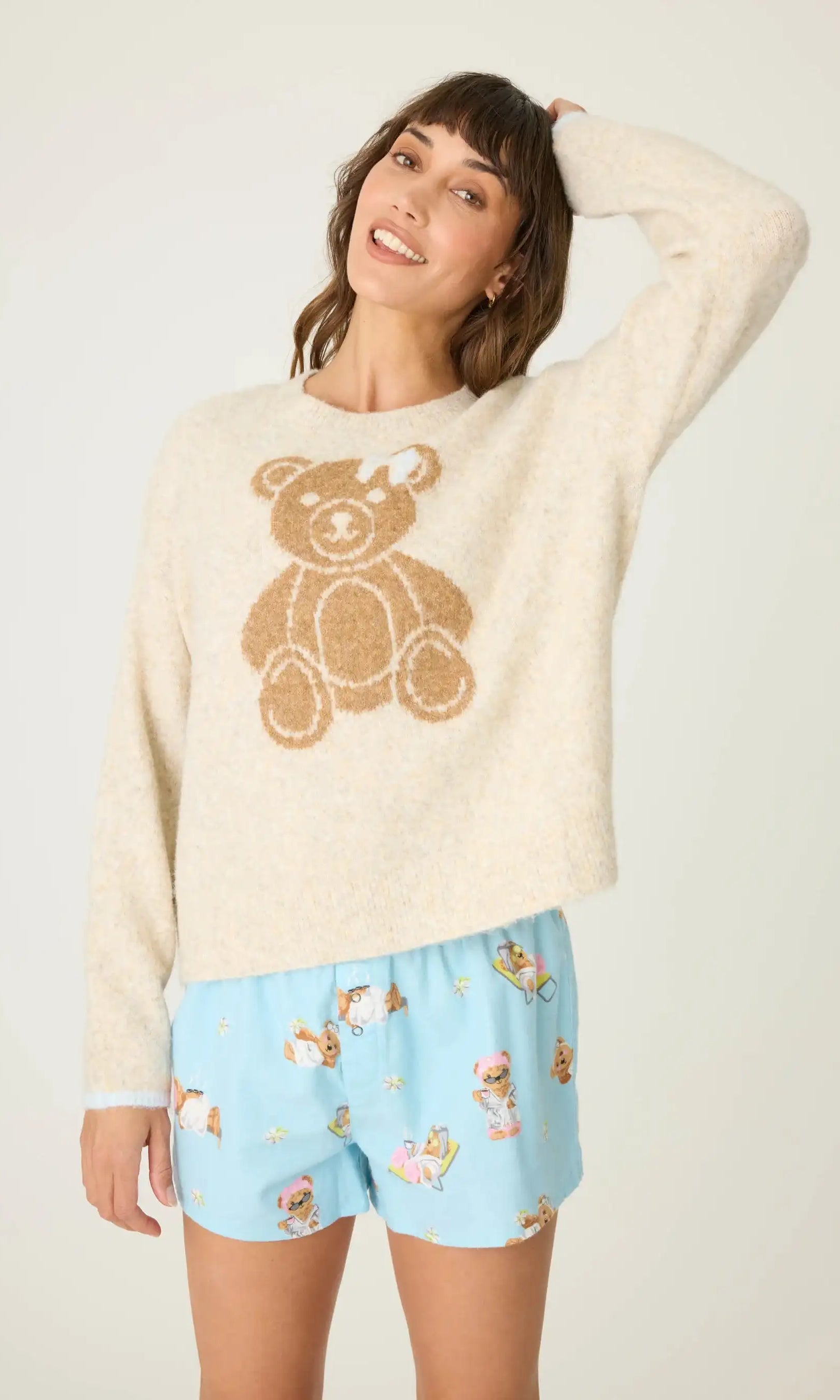Woman wearing a cream sweater with a teddy bear design and light blue shorts with teddy bear pattern on a white background