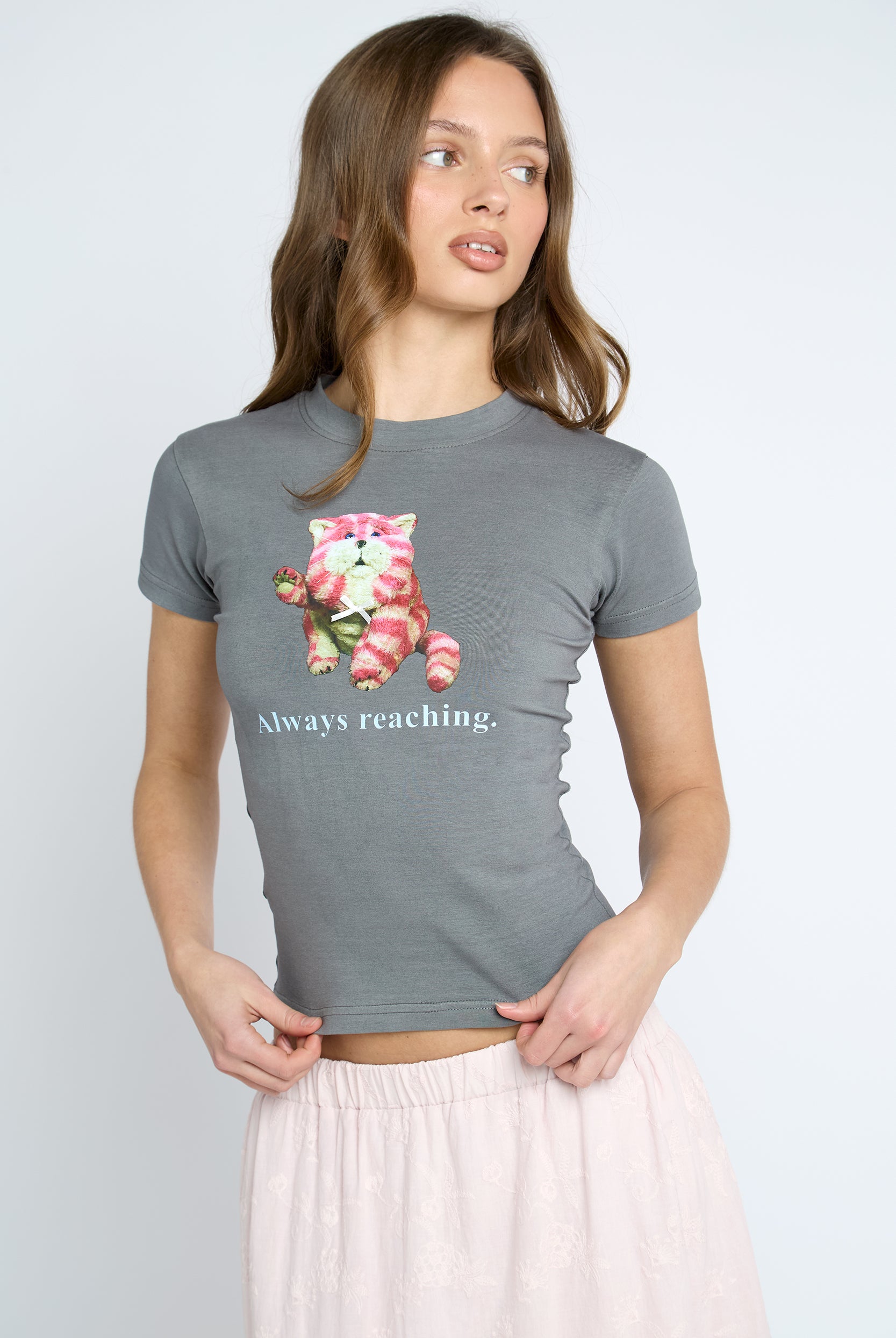 A woman wearing a heather gray short-sleeve t-shirt with a graphic of a pink and white cat holding a strawberry and the text 'Always reaching.' below it, paired with a light pink skirt, against a light gray studio background.