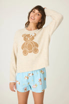 Woman wearing a cream sweater with a teddy bear design and light blue shorts with teddy bear pattern on a white background